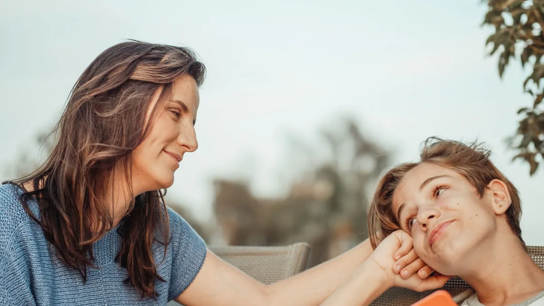 A parent leaning in toward her teenage son, her hand resting gently at his cheek as they smile at each other.