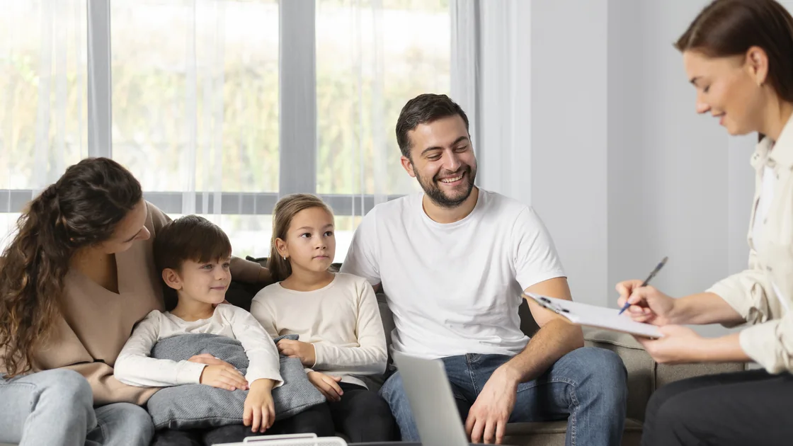 A family of four sitting together on a sofa, talking with a therapist who is taking notes beside them.