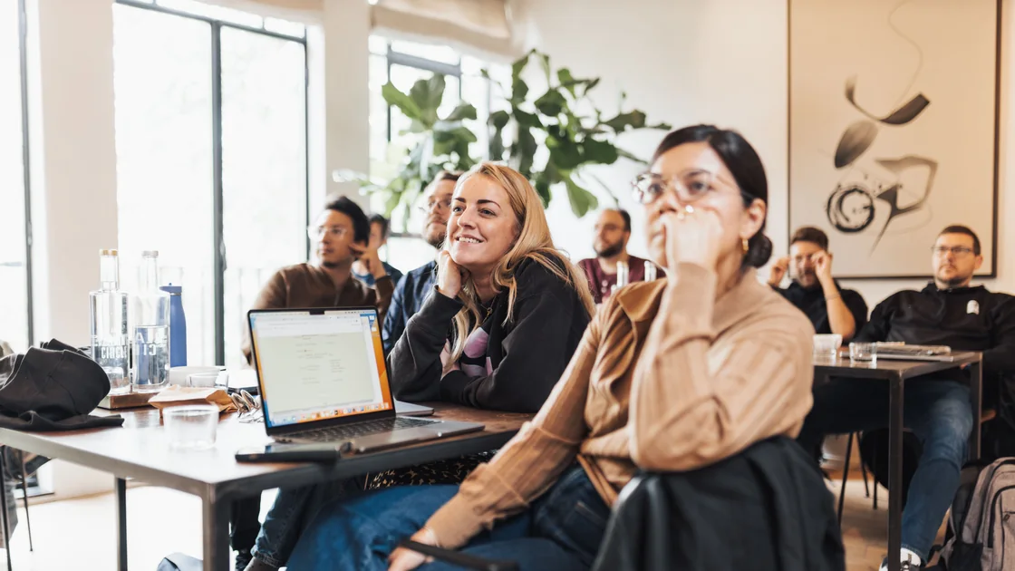 A group of people seated around tables in a bright room, listening attentively to a workshop presentation.