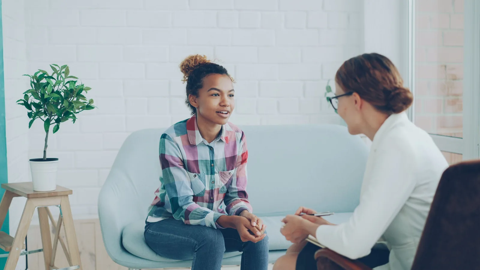 A young adult talking with her therapist in a bright room, the therapist taking notes beside her.