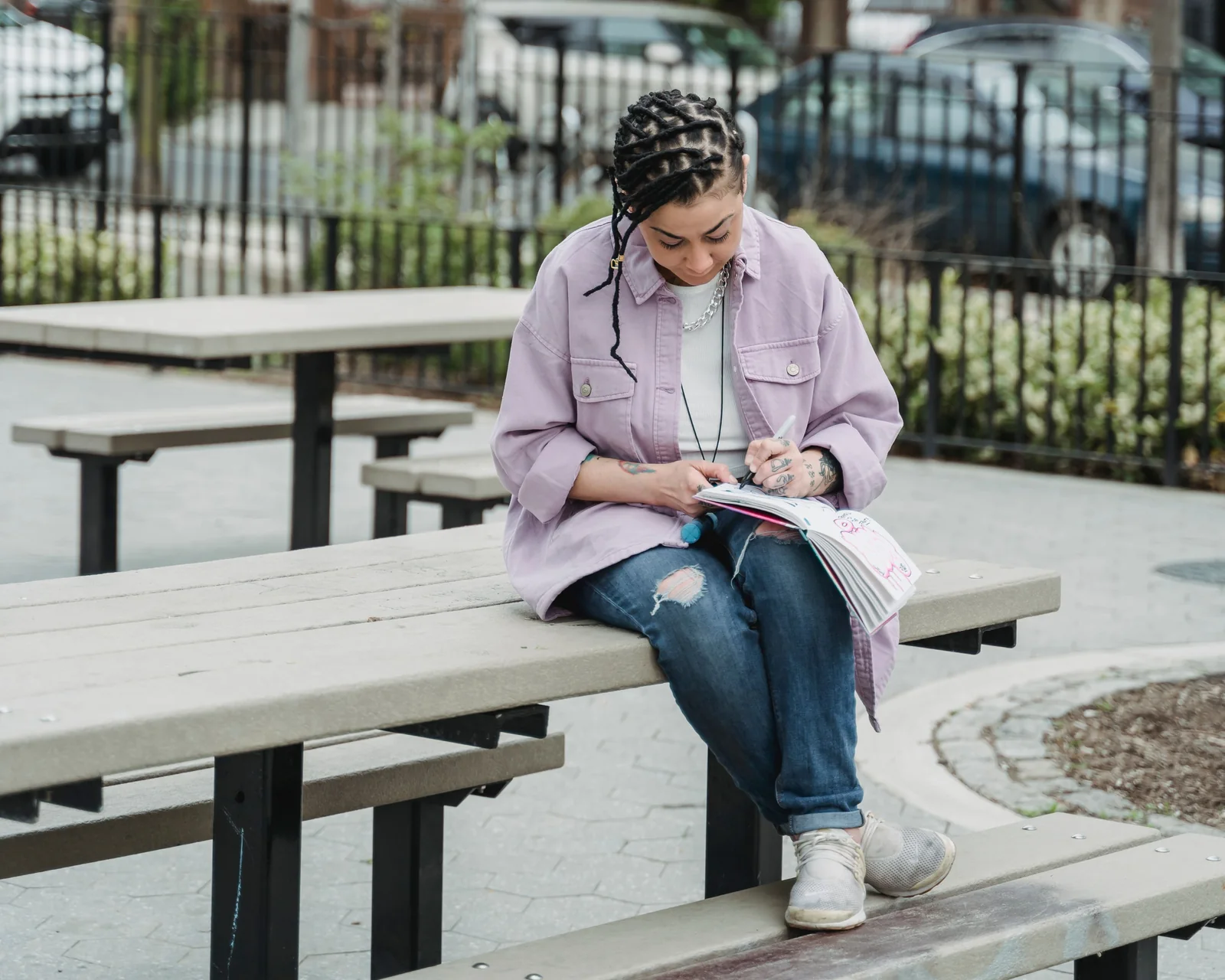 A young person sitting cross-legged on a picnic table outside, drawing in a notebook.
