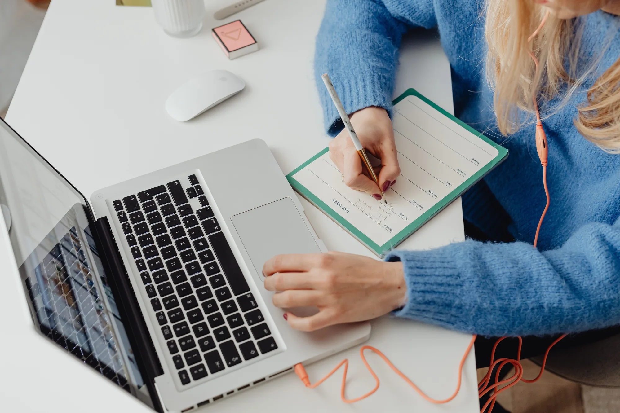 A woman sitting at a desk with a laptop open, writing this week's plan into a paper planner, earphones in.