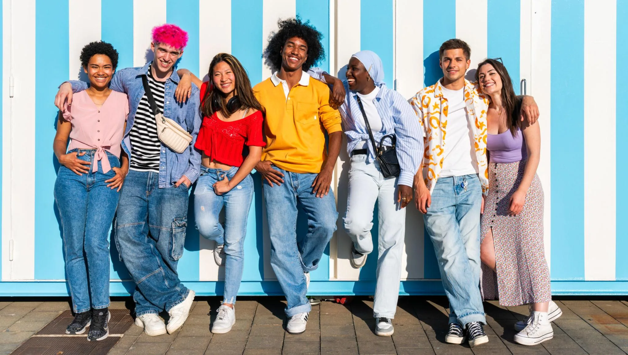 Seven young adults standing together against a blue and white striped wall, arms around one another, laughing.