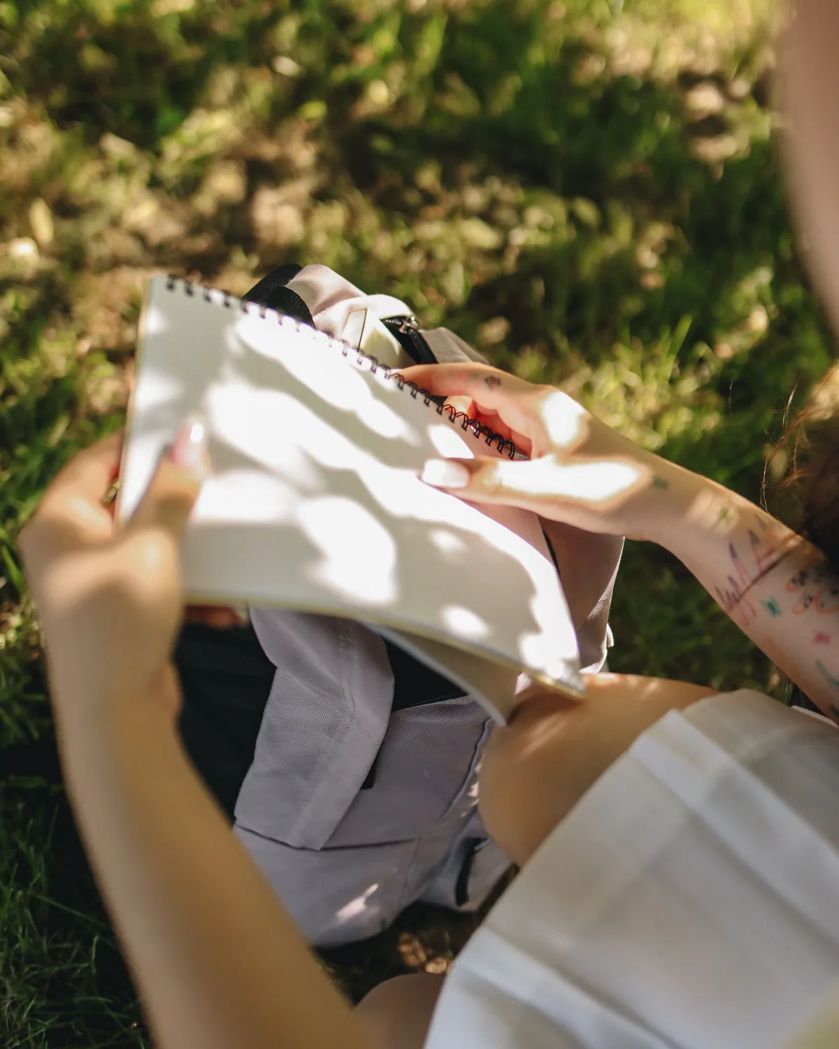 Photographed from above, a person seated on grass holding a blank spiral sketchbook open on their lap, a grey backpack resting between their knees and dappled leaf shadows falling across the empty pages.