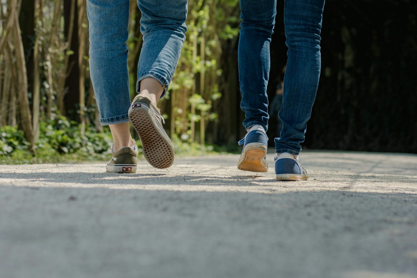 Two people walking side by side on a sunlit path under green trees, seen from behind; one foot is mid-stride, lifting off the ground.